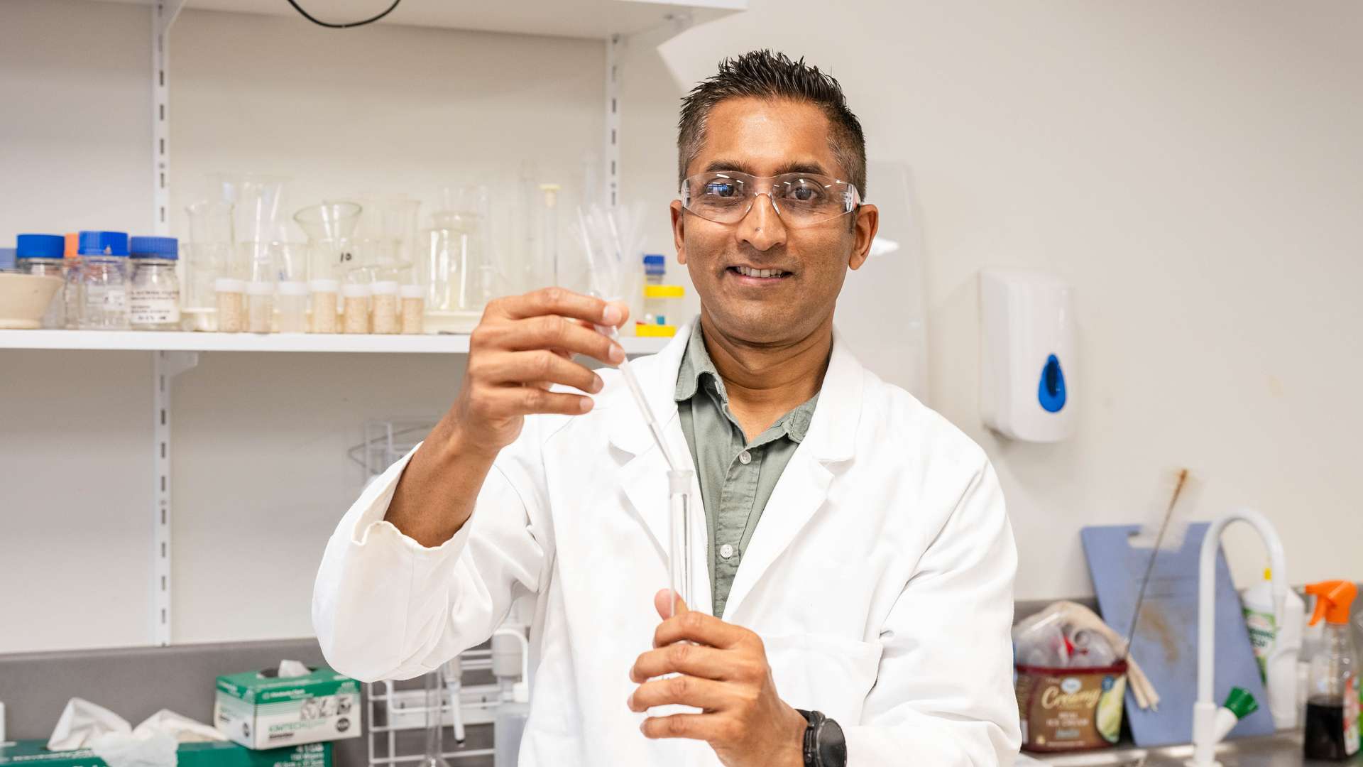 Dr Shaneel Chandra wears a white lab coat and stands in a laboratory.