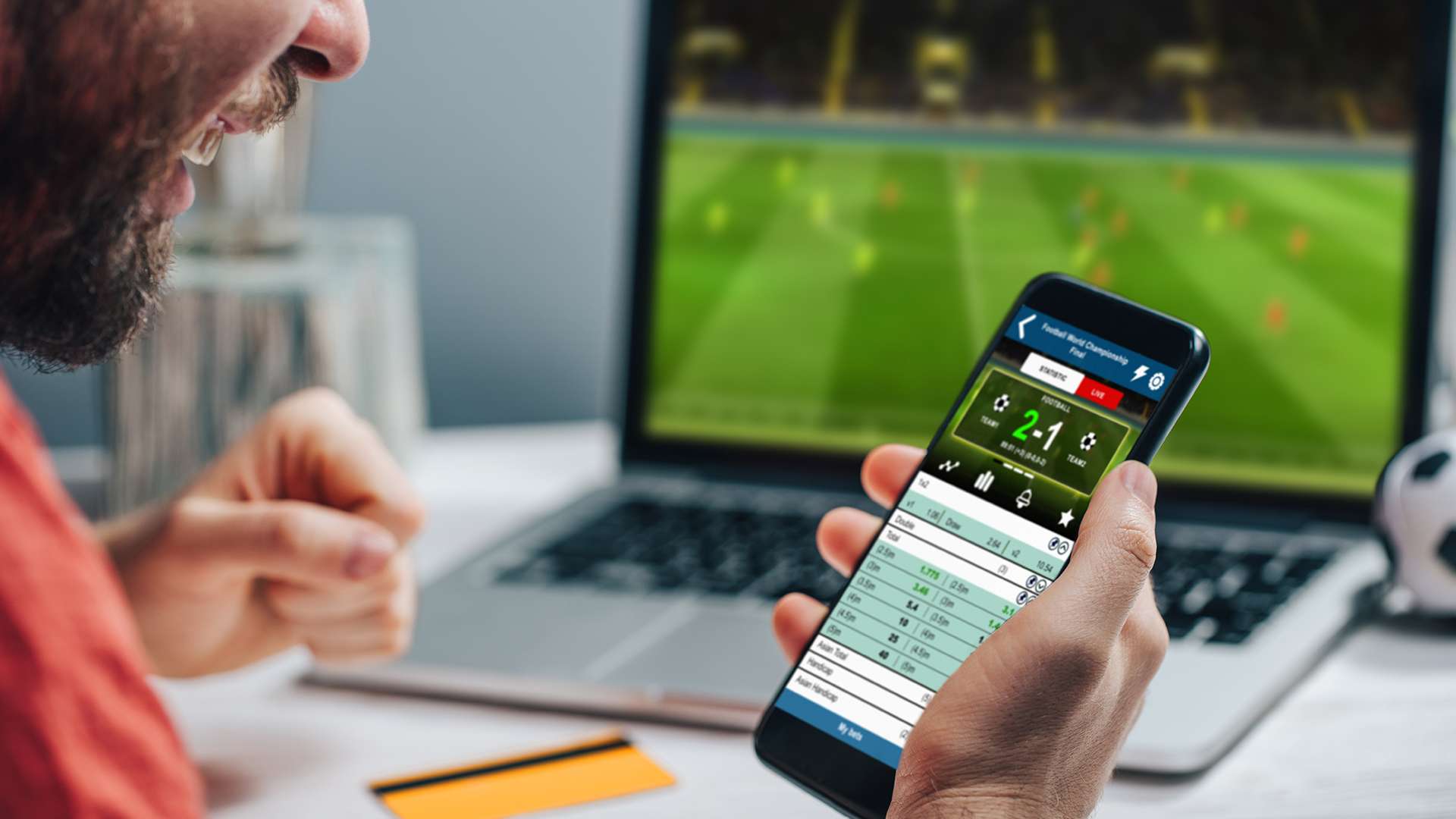 Person eagerly looking at their phone with a gambling app open as a soccer game plays on a laptop in the background