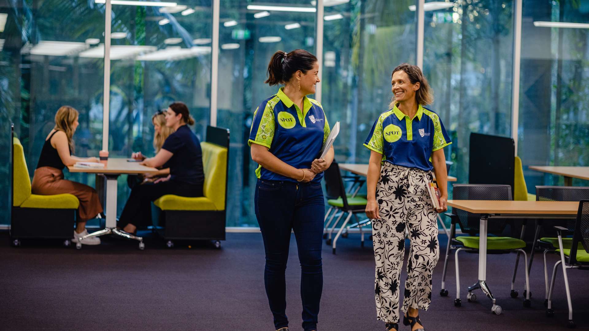 Two CQU staff walking in library smiling at each other wearing SPOT shirts.