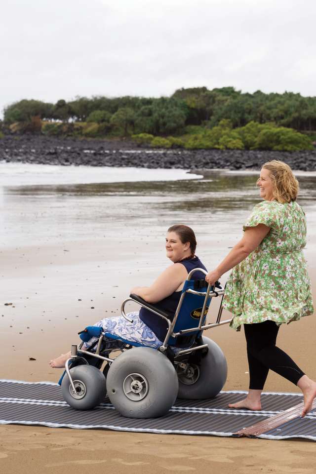 Student helping another student in a wheelchair down the beach smiling at the ocean.