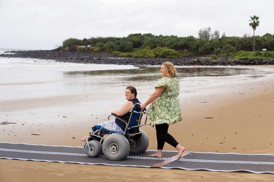 Student helping another student in a wheelchair down the beach smiling at the ocean.